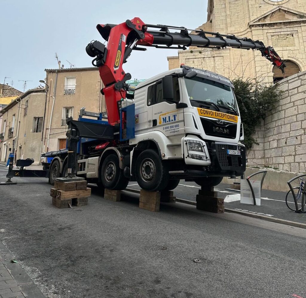 Camion-grue M.L.T Palfinger : Levage exceptionnel en ville Camion-grue blanc M.L.T Palfinger sur cales en bois, CONVOI EXCEPTIONNEL, avec sa grue rouge étendue vers un bâtiment historique à horloge.