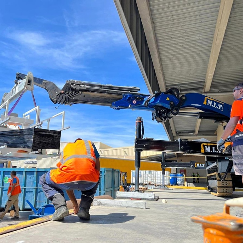 Grue M.L.T. Palfinger bleue levant une palette de matériaux sur un site industriel. Deux ouvriers en orange supervisent la manutention sous ciel bleu.