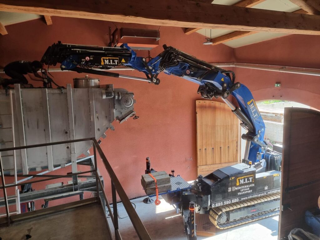 Mini-grue M.L.T. bleue et noire installant une grande cuve métallique dans un bâtiment aux murs rouges. Un homme travaille sur la cuve.