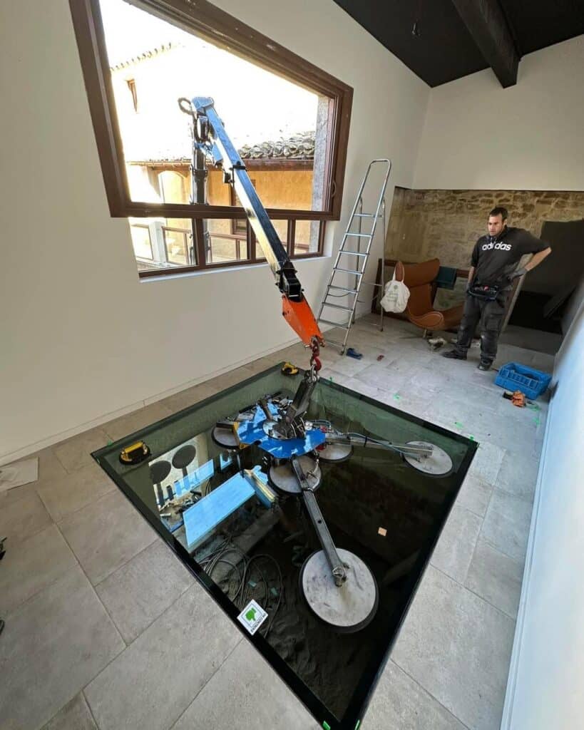 Installation plancher vitré : pose dalle de verre par grue Grue robotisée positionnant une grande dalle de verre EN CONTROL pour un plancher vitré. Un homme en t-shirt Adidas supervise l'installation.