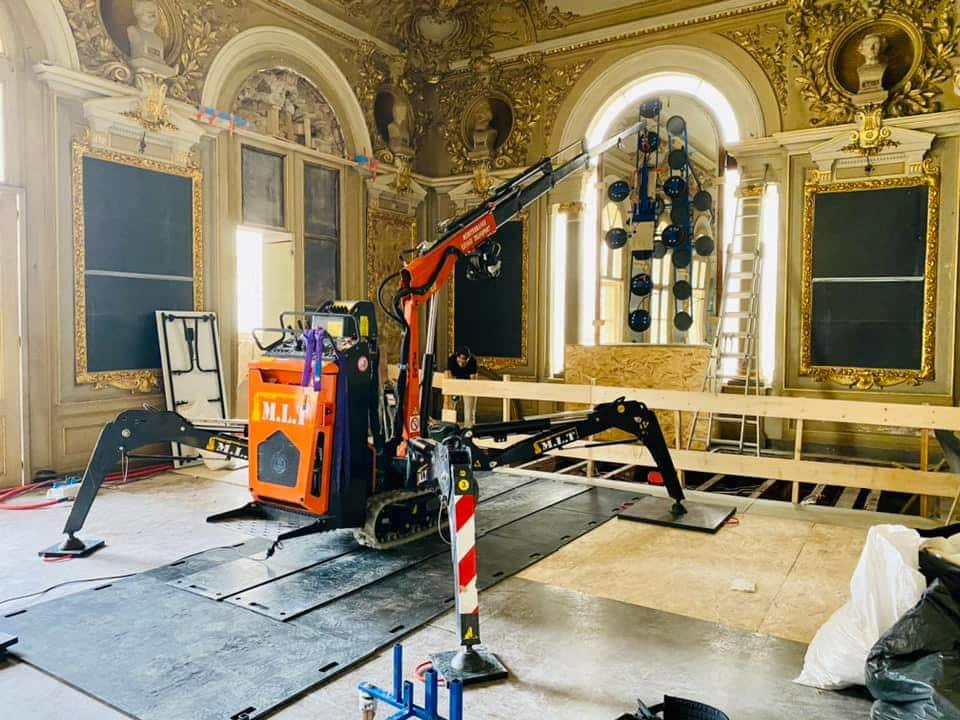 Installation vitrage grand format : mini-grue MLT en salle historique Mini-grue M.L.T. sur chenilles installant une grande vitre avec système à ventouses dans une salle dorée et ornementée d'un bâtiment historique.
