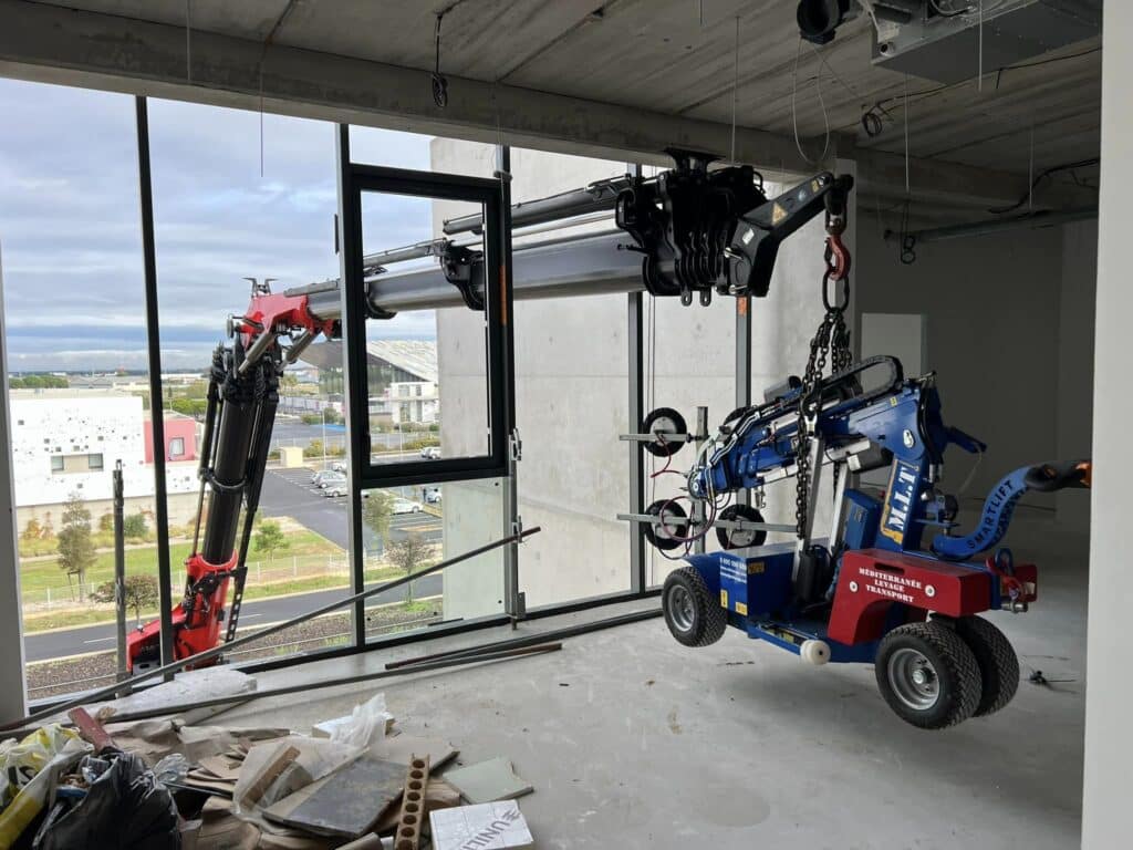 Installation de vitre avec grue et lève-vitre Smartlift Un bras de grue installe une grande vitre avec un lève-vitre robotisé "M.L.T. SMARTLIFT MÉDITERRANÉE LEVAGE TRANSPORT" sur chantier.