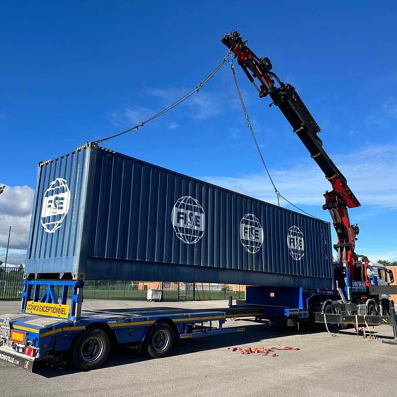 Levage container FISE par camion grue – Convoi Exceptionnel Container FISE bleu levé par camion-grue rouge sur une remorque surbaissée "CONVOI EXCEPTIONNEL" sous ciel bleu.