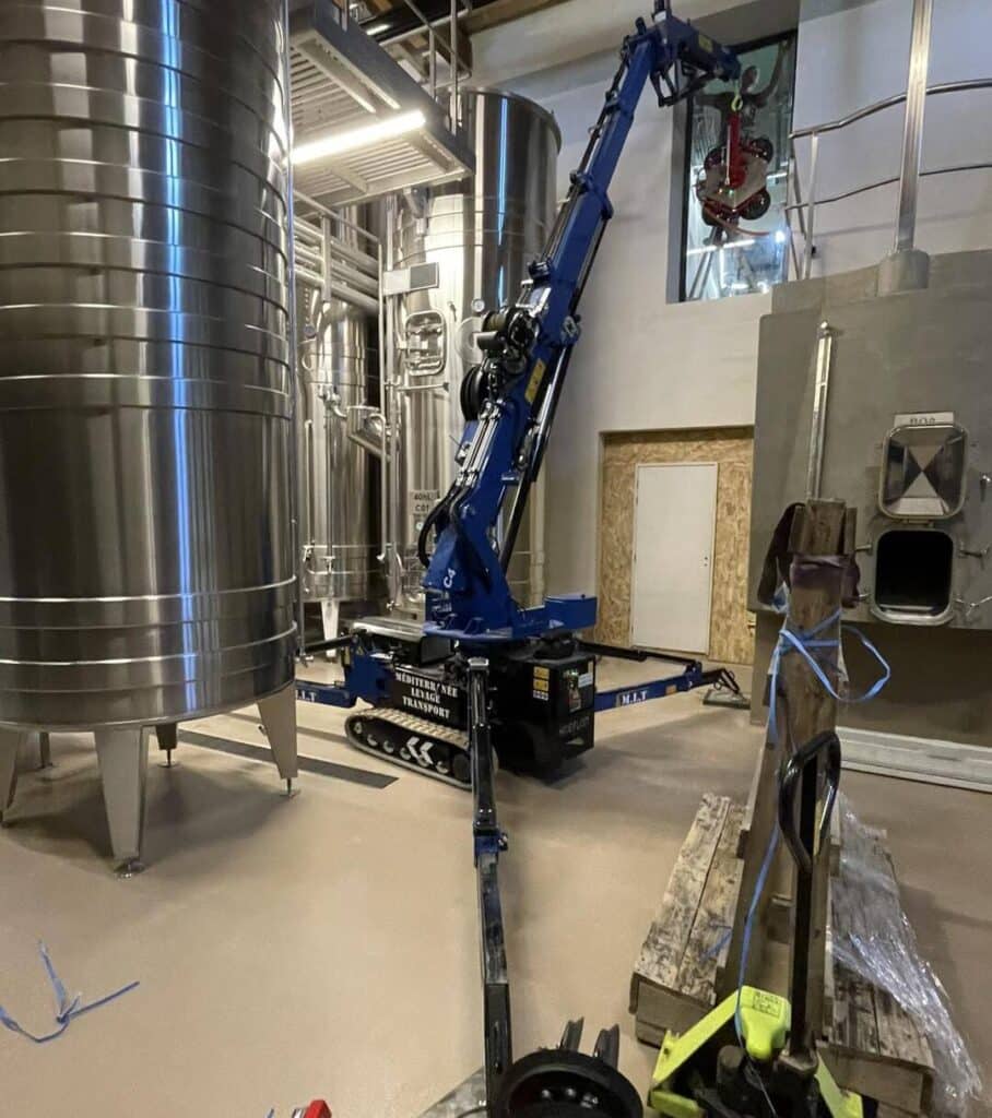 Mini-grue Hoeflon bleue (MEDITERRANEE LEVAGE TRANSPORT) lève un équipement rouge au milieu de grandes cuves inox dans un entrepôt industriel.