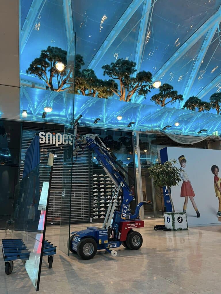 Machine de levage MLT installant un grand panneau de verre devant le magasin Snipes. Plafond bleu avec arbres lumineux et pub pour enfants.