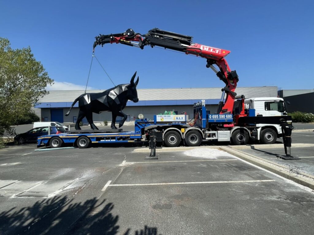 Grue M.L.T. (0 695 590 680) soulevant une sculpture de taureau polygonal noir d'un camion-remorque bleu, sous un ciel clair.