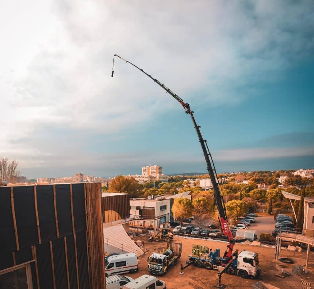 Camion-grue TMT au bras déployé sur un chantier de construction. La flèche s'élève au-dessus des structures en bois et du paysage urbain.