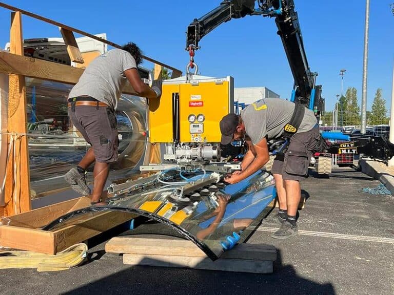 Deux professionnels manipulent un grand vitrage incurvé. Le levage est effectué par une ventouse fixée à une mini-grue M.L.T.