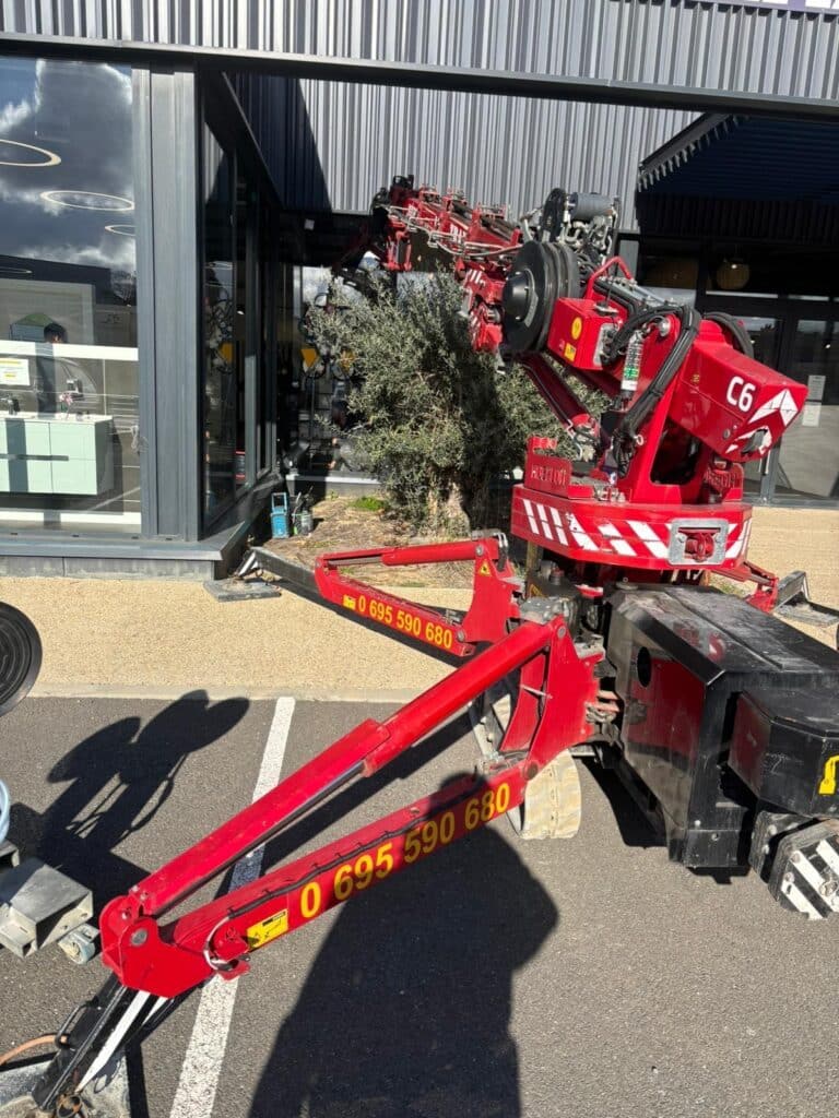 Mini grue araignée rouge C6 sur chenilles, outriggers déployés pour travaux devant façade vitrée. Contact: 0 695 590 680.