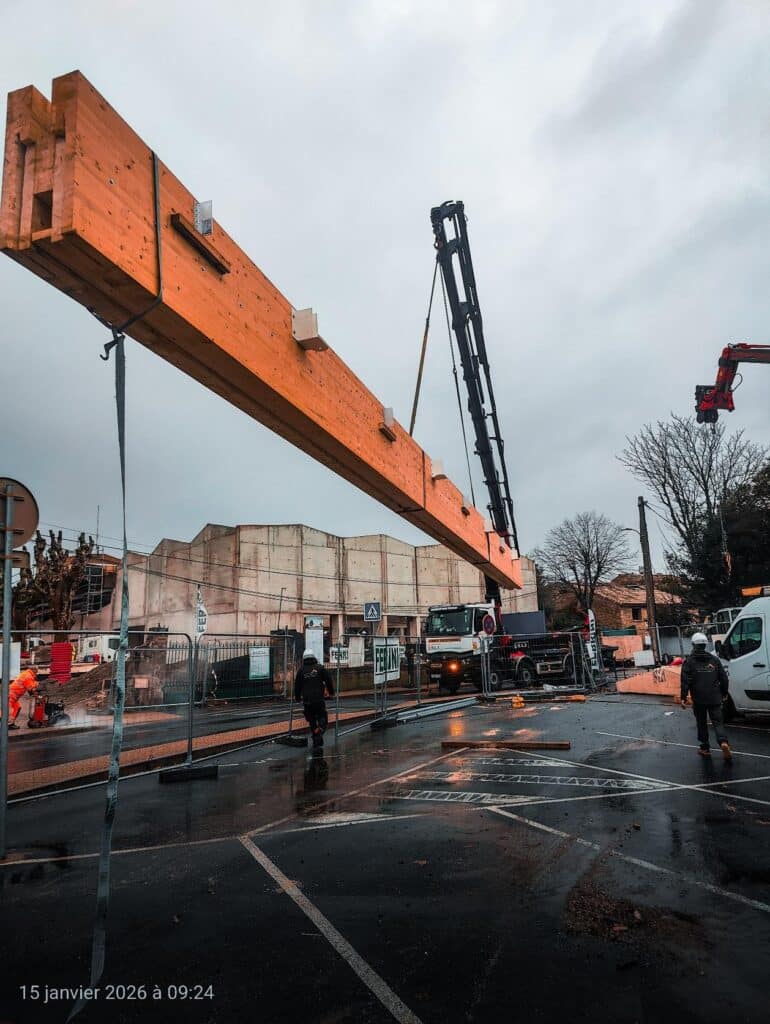 Levage d'une énorme poutre en bois lamellé-collé par grue sur un chantier mouillé. Ouvriers présents. 15 janvier 2026 à 09:24.