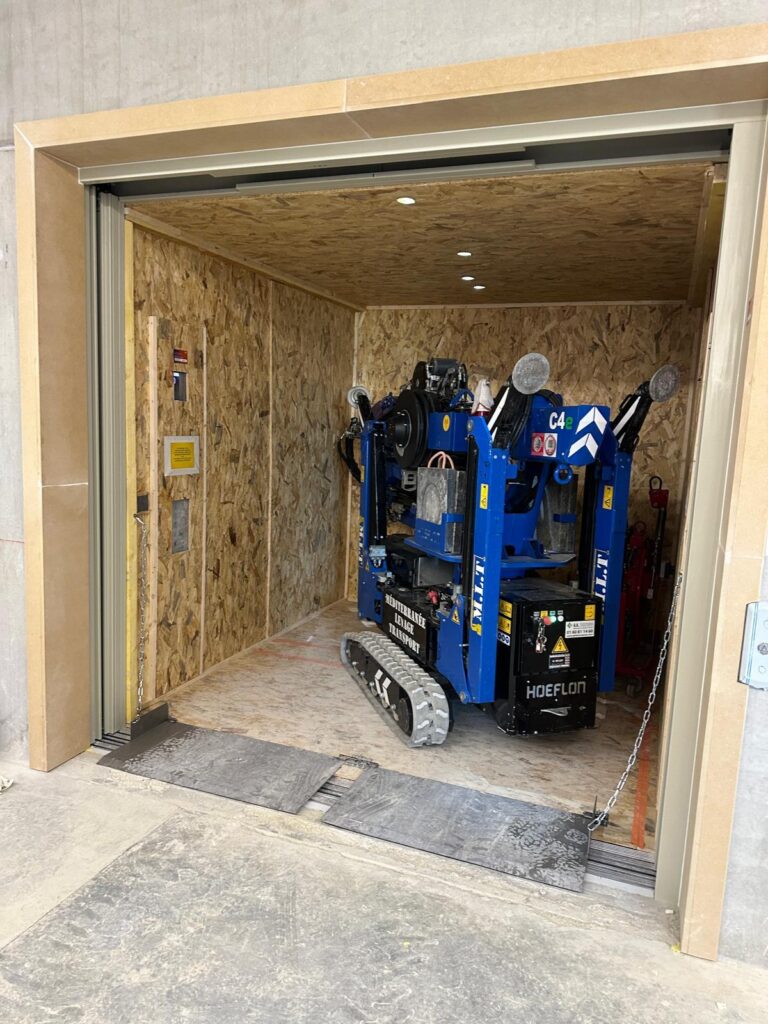 Robot de levage M.L.T. HOEFLON C4e bleu sur chenilles stationné dans un monte-charge de chantier provisoire en OSB avec rampes d'accès.