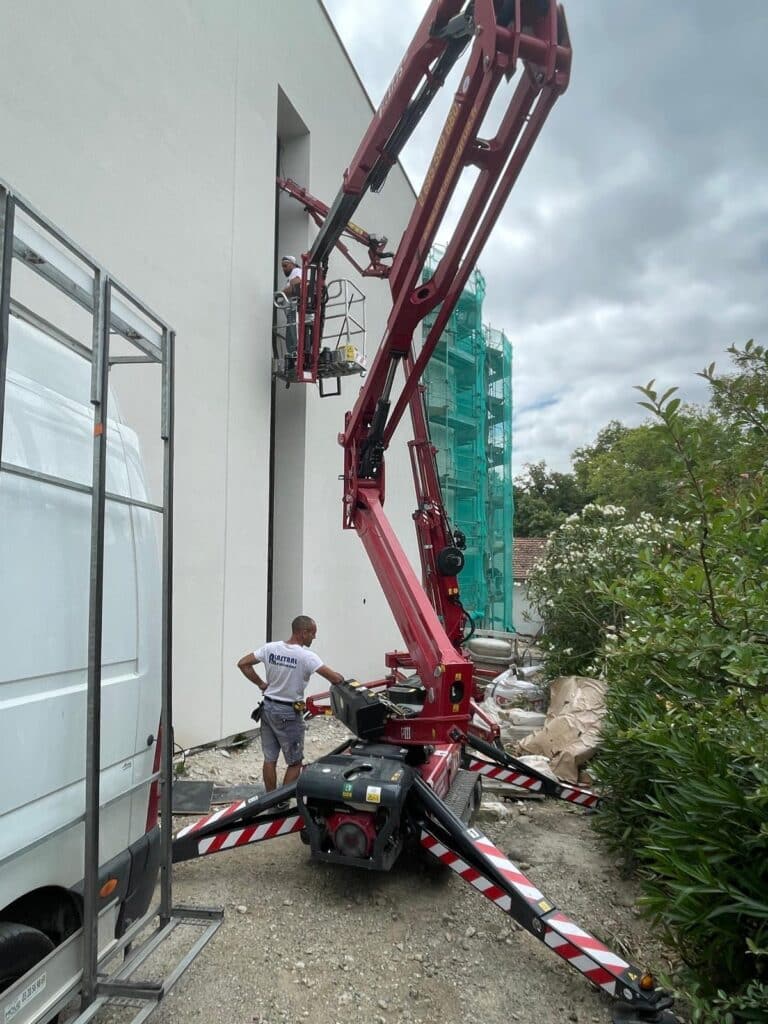 Deux ouvriers réalisent des travaux sur une façade blanche à l'aide d'une nacelle élévatrice rouge (mini-grue). Un ouvrier au sol porte un T-shirt PLASTRAL.